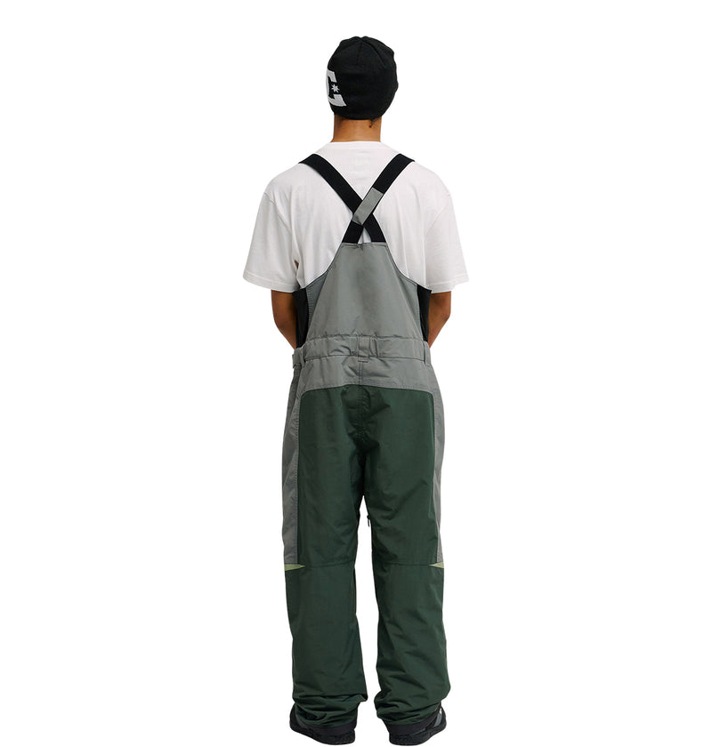 A person facing away, wearing a black beanie, white T-shirt, and DC Shoes Mountain Lab Bib 20K two-tone green overalls with black straps designed for breathable protection, standing against a plain white background.