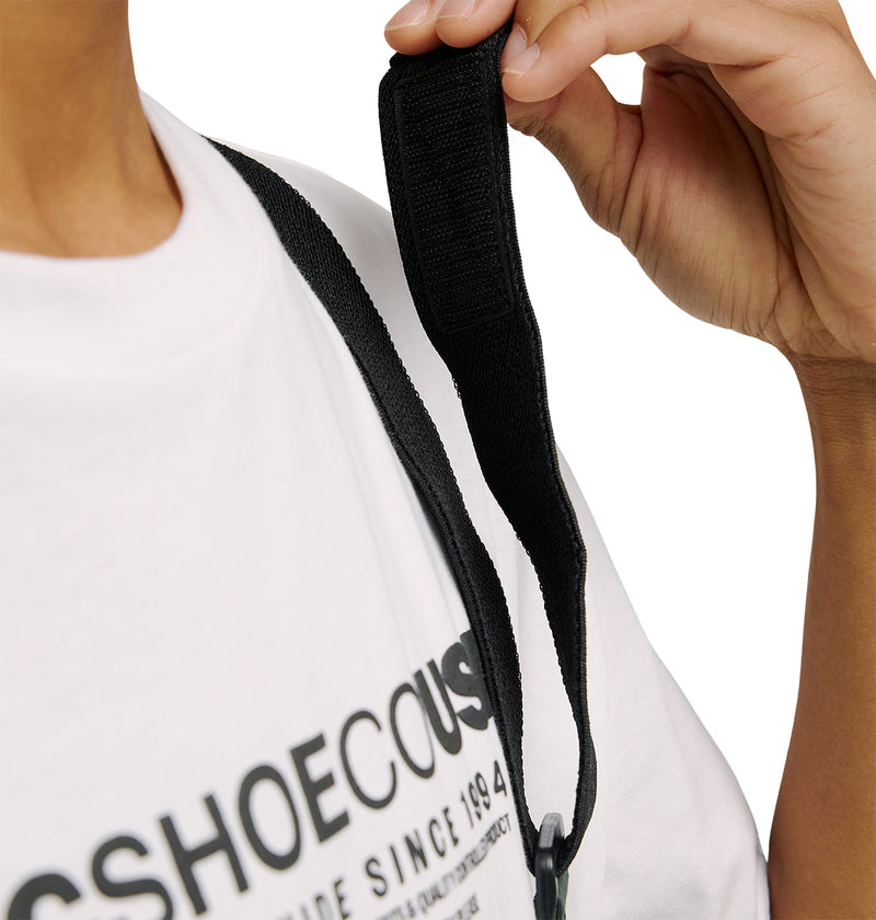 A person wearing a white T-shirt with black printed text adjusts the black strap of a bag on their shoulder while dressed in the DC Shoes Mountain Lab Bib 20K, which offers breathable protection. Only part of their upper body and one hand are visible.