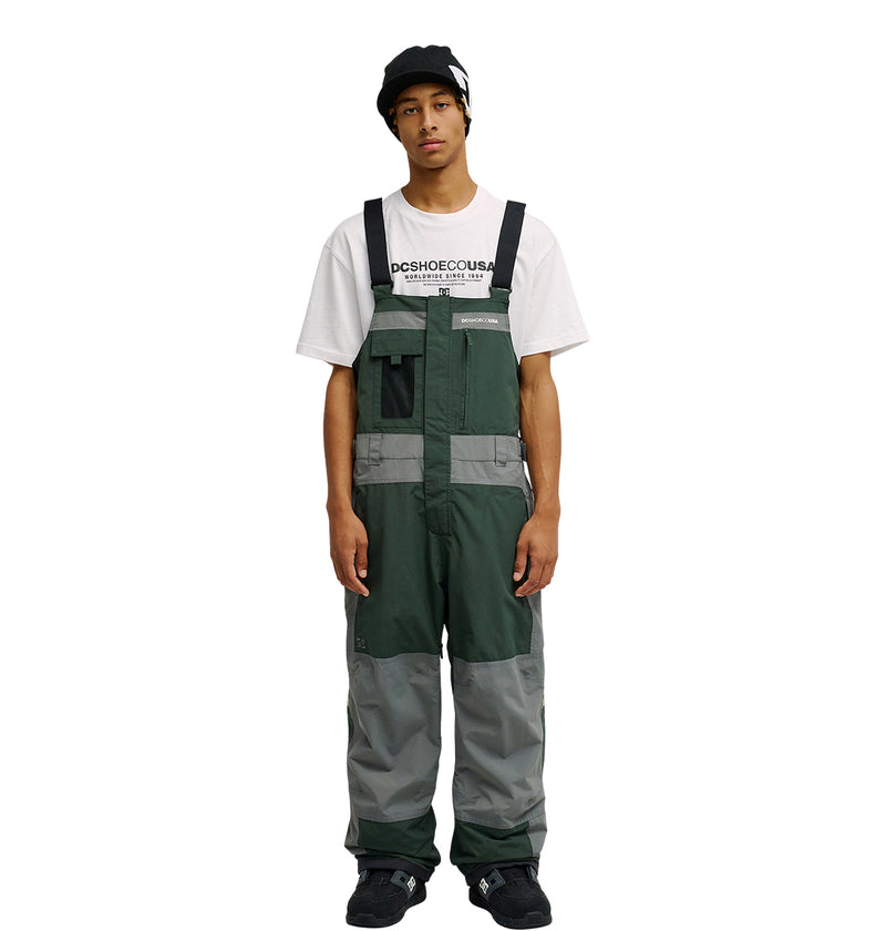 Wearing the DC Shoes Mountain Lab Bib 20K in green and gray, along with a black beanie and a white graphic T-shirt, a person stands facing forward with arms relaxed at their sides against a plain white background.