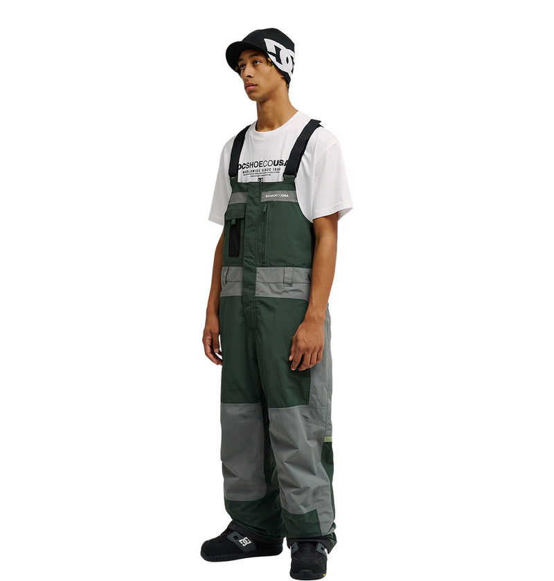 A person wearing a black and white beanie, a white T-shirt, and the green and gray DC Shoes Mountain Lab Bib 20K stands facing left with a neutral expression against a plain white background.