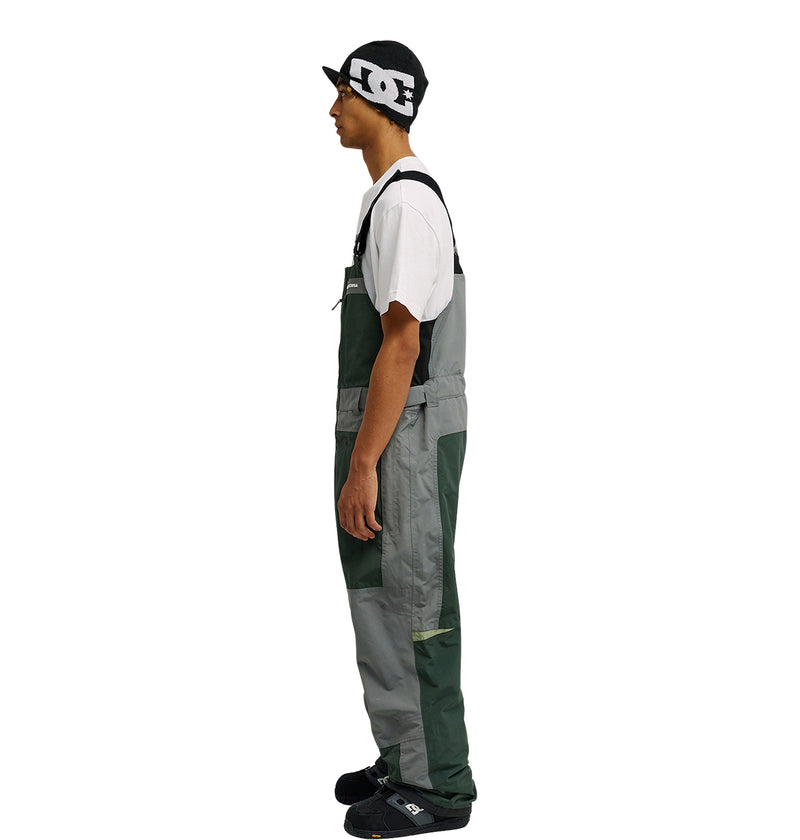 A person stands in profile against a plain white background, wearing the DC Shoes Mountain Lab Bib 20K—gray-green overalls paired with a black beanie, white t-shirt, and black shoes—demonstrating its breathable protection for all-day comfort.