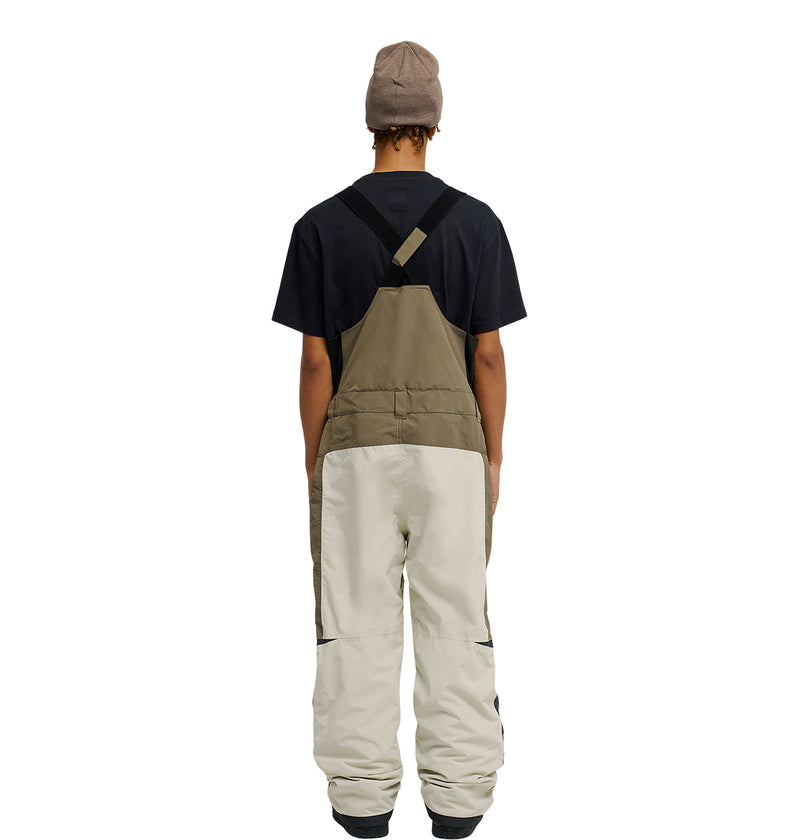A person wearing a black T-shirt, the DC Shoes Mountain Lab Bib 20K two-tone overalls (brown top, beige bottom) designed for breathable protection, and a beige beanie stands facing away from the camera against a white background.