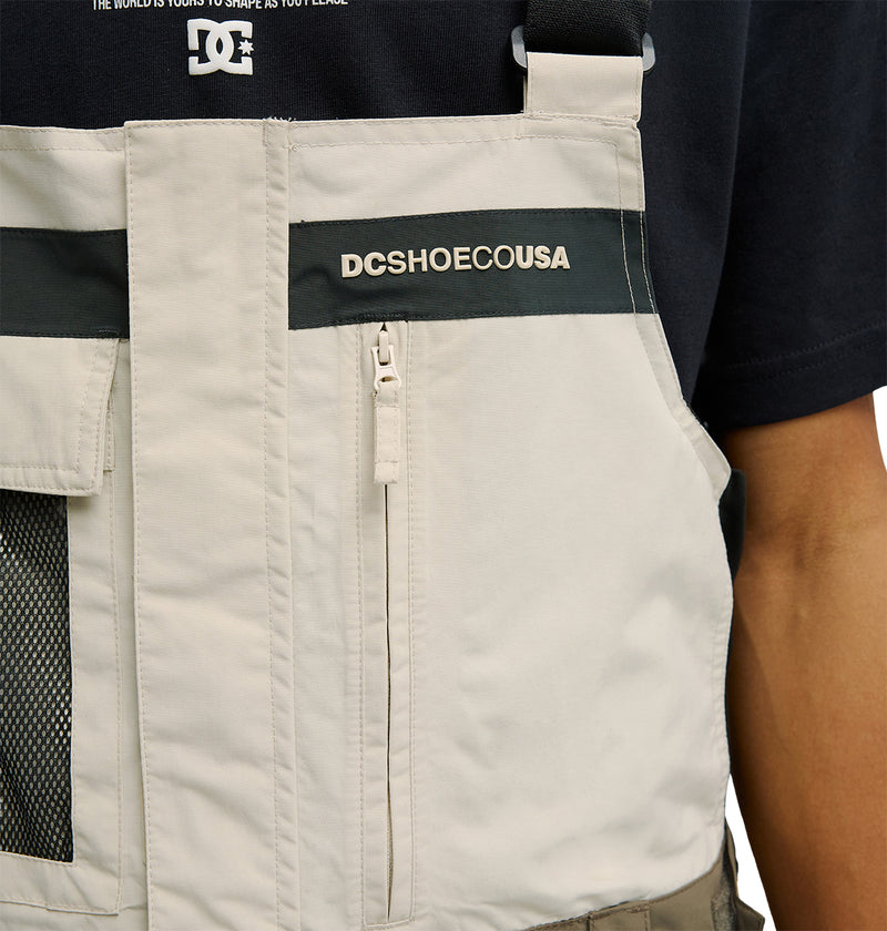 Close-up of a person wearing the DC Shoes Mountain Lab Bib 20K—a beige vest featuring a zippered pocket and DCSHOECOUSA logo—over a black shirt. Only the upper torso is visible, emphasizing the bib’s breathable protection for optimal comfort.