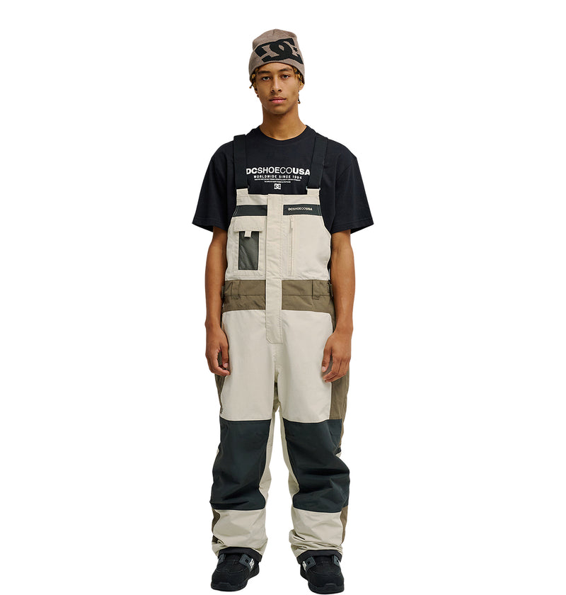 A person wearing a beanie, a black DC Shoes t-shirt, and the Mountain Lab Bib 20K bib overalls in beige, brown, and gray stands facing forward with a neutral expression. The DC Shoes Mountain Lab Bib 20K is designed for reliable snow performance, offering both style and functionality on colder days.