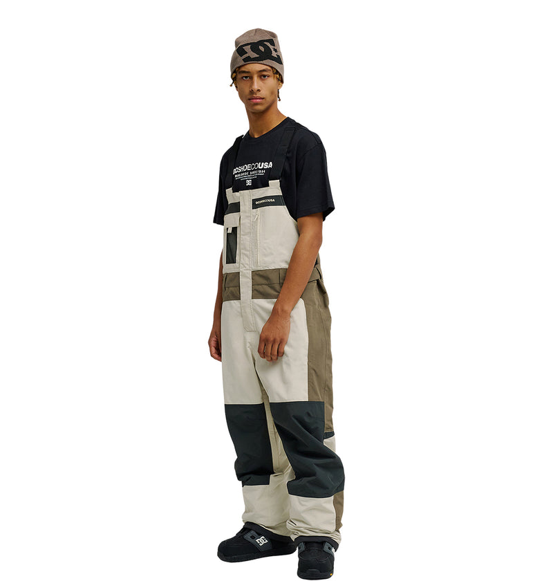 Wearing a graphic T-shirt, the individual is styled in DC Shoes’ Mountain Lab Bib 20K color-blocked overalls in beige, tan, and gray—designed for breathable protection—paired with black shoes and a brown beanie, standing slightly turned against a plain white backdrop.