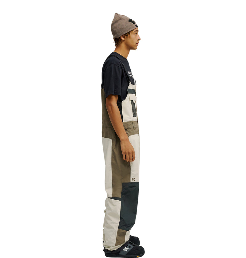 A person in profile view wears the DC Shoes Mountain Lab Bib 20K overalls, designed for breathable protection, paired with a black T-shirt, black shoes, and a taupe beanie. The background is plain white.
