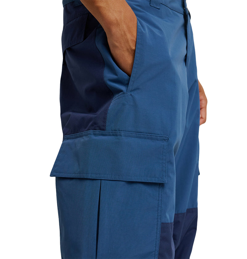 Wearing the DC Shoes Mountain Lab Pants 10K, a person stands with their right hand in the pocket. These waterproof pants showcase a large flap pocket on the thigh and feature dark blue panels running along the side and inside of the leg.