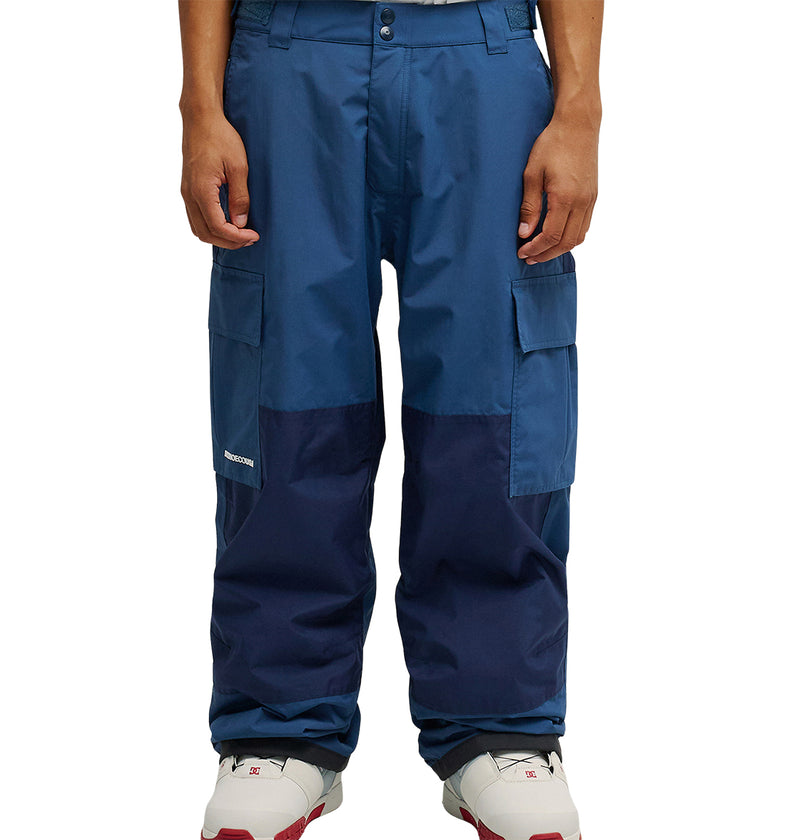 A person wearing DC Shoes’ Mountain Lab Pants 10K in blue—with darker blue knee patches—and white sneakers stands against a white background. These cargo pants from DC Shoes offer breathable protection, ensuring all-day comfort.