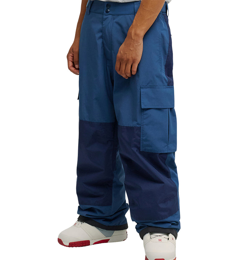 A person is wearing DC Shoes’ Mountain Lab Pants 10K—loose-fitting, breathable cargo pants in two shades of blue—paired with white sneakers featuring red accents. Only the pants and lower legs are visible against a plain white background.