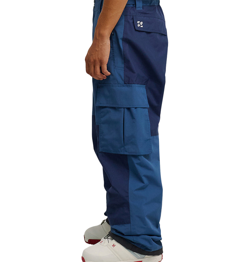 A person wearing DC Shoes Mountain Lab Pants 10K in blue, featuring large side pockets and white footwear, is photographed in profile against a plain white background. The image focuses on the lower half of the body, showcasing pants engineered by DC Shoes for breathable protection and all-day comfort in any environment.