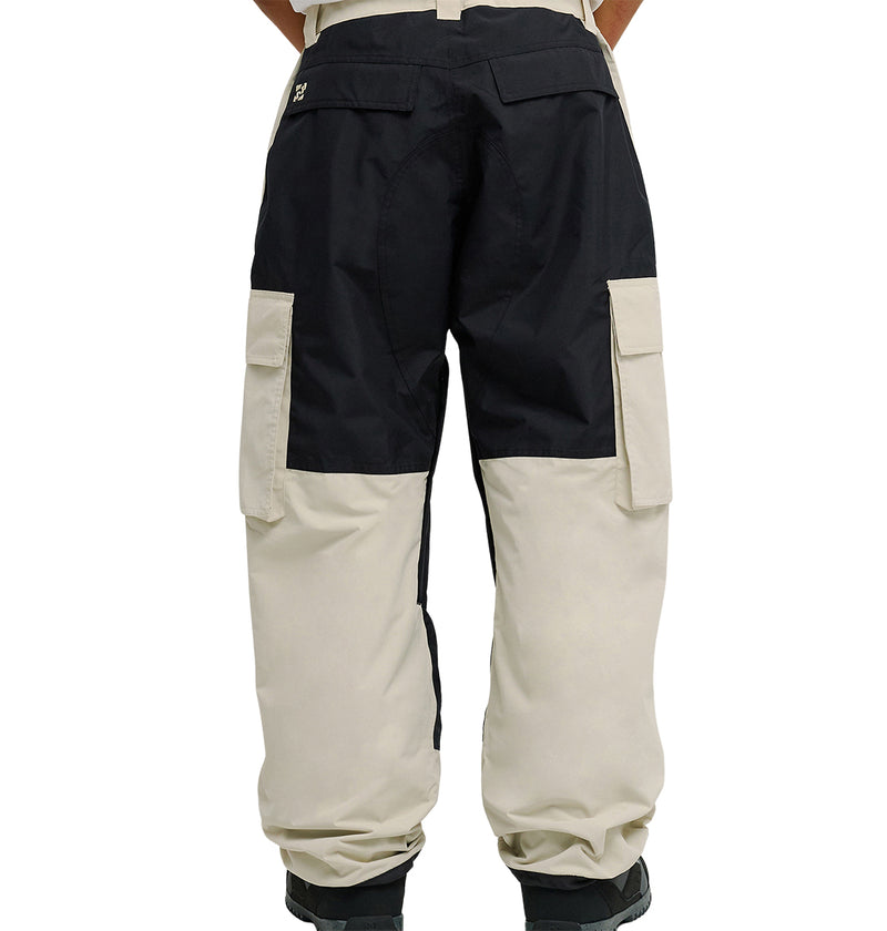 Wearing DC Shoes’ Mountain Lab Pants 10K, which showcase a two-tone design with black on the upper section and beige on the lower section, a person stands with their back to the camera. These pants deliver both standout style and snow-ready performance, featuring large side pockets, while black boots are partially visible.