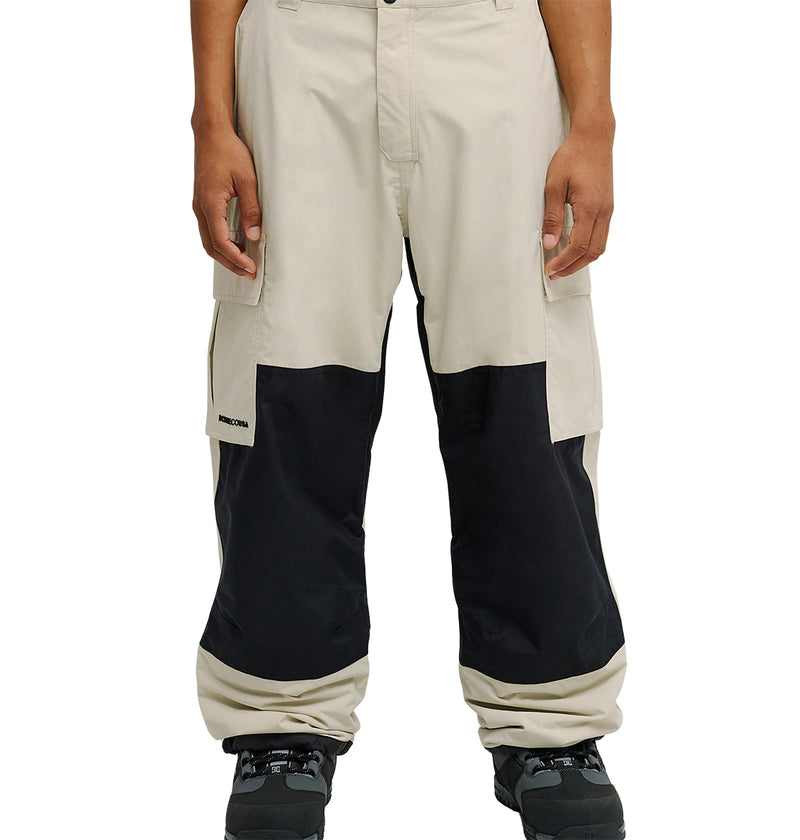 A person is wearing the DC Shoes Mountain Lab Pants 10K in beige, featuring large black patches on the knees and lower legs for breathable protection. These loose-fitting pants are shown from the waist down and paired with black shoes.