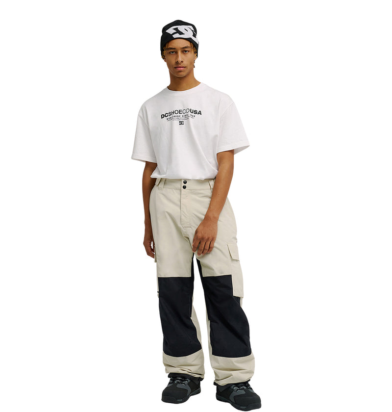 Wearing a black-and-white headband, a white DC SHOES USA t-shirt, Mountain Lab Pants 10K by DC Shoes—beige cargo pants with black knee patches featuring breathable protection—and black sneakers, the person stands against a plain white background.