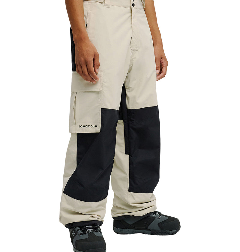 A person wearing the Mountain Lab Pants 10K by DC Shoes in beige and black, paired with black boots, stands with hands relaxed at their sides. The pants feature black knee patches and a side pocket with a small logo, offering breathable protection designed for enhanced snow performance.