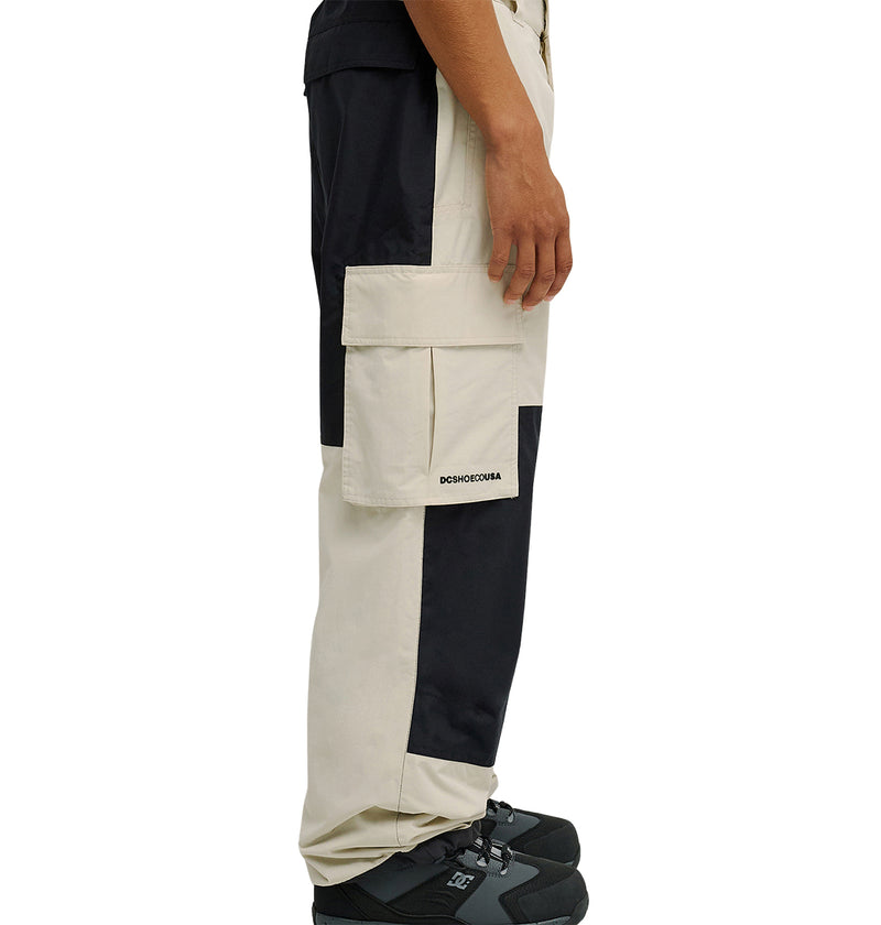 A person wearing DC Shoes’ Mountain Lab Pants 10K in a black and white color-block design and black sneakers stands sideways, displaying the pants’ side pocket and signature DSCHDSCHDSCHA text, highlighting the breathable protection provided by these pants.