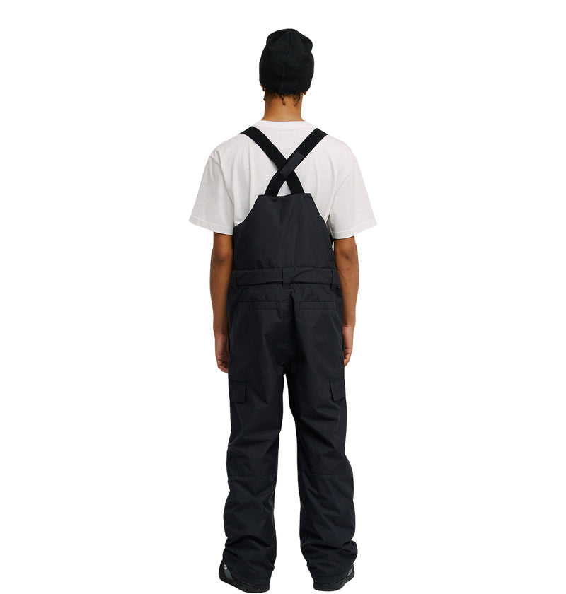 A person stands facing away against a plain white background, wearing a white t-shirt, black shoes, a black beanie, and the DC Shoes Docile Bib 10K—black insulated bib overalls featuring crisscross straps on the back.