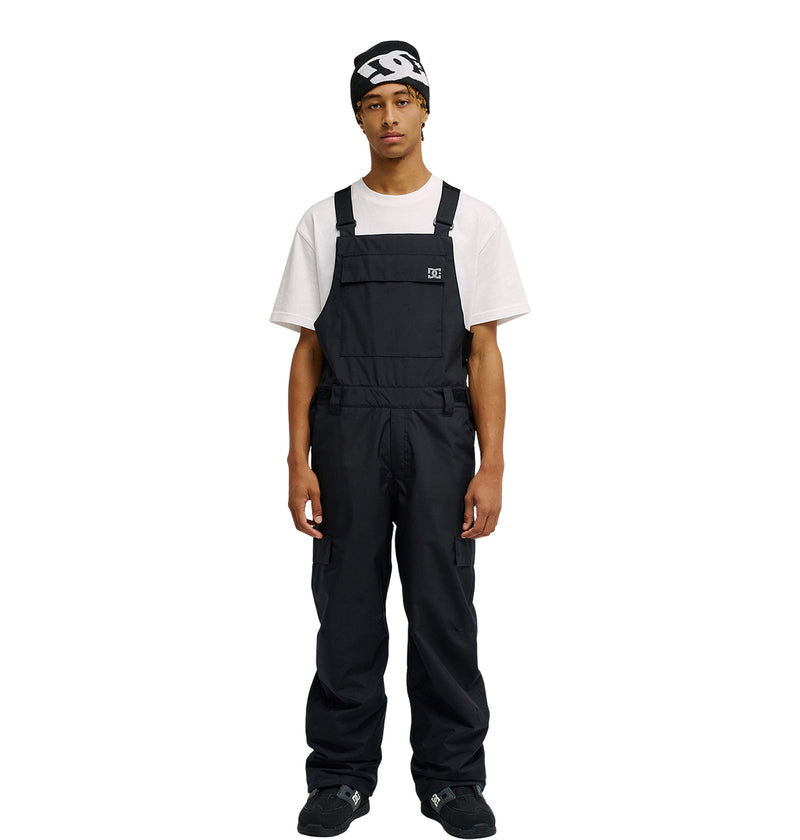 A person stands facing forward against a plain white background, wearing the DC Shoes Docile Bib 10K black snow bib overalls over a white T-shirt, paired with black shoes and a black beanie featuring a white logo.