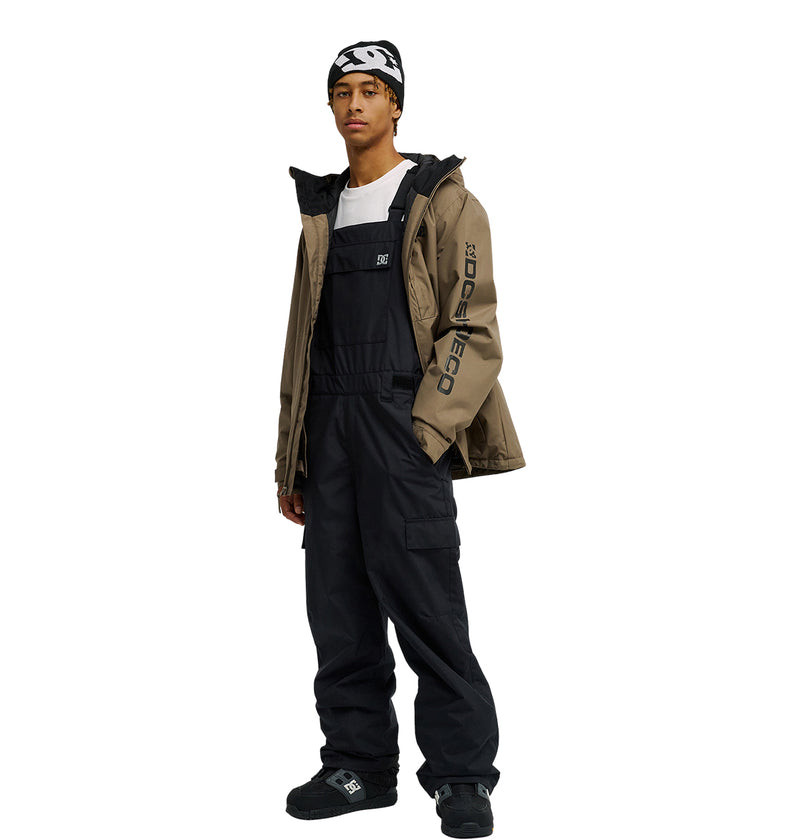 A person wearing a beanie, tan jacket with “Dope” on the arm, white shirt, black shoes, and the DC Shoes Docile Bib 10K black insulated overalls stands against a white background.