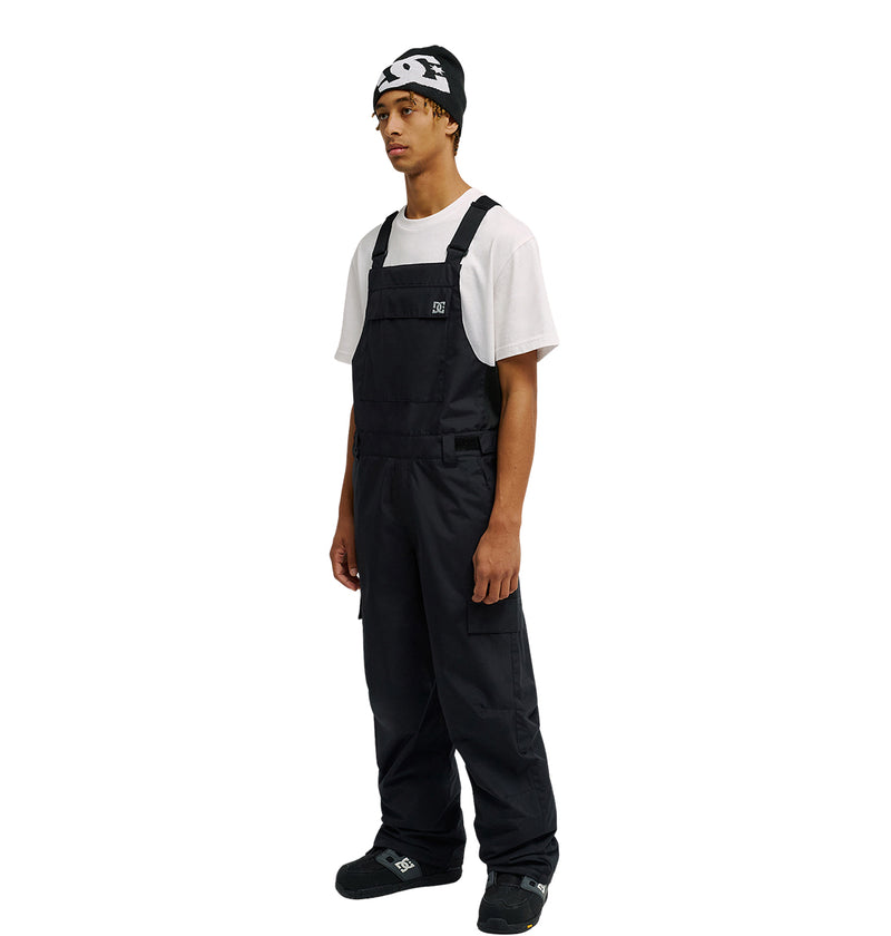 A person wearing a white t-shirt, black DC Shoes Docile Bib 10K snow overalls, black shoes, and a black beanie with a white logo stands against a plain white background.