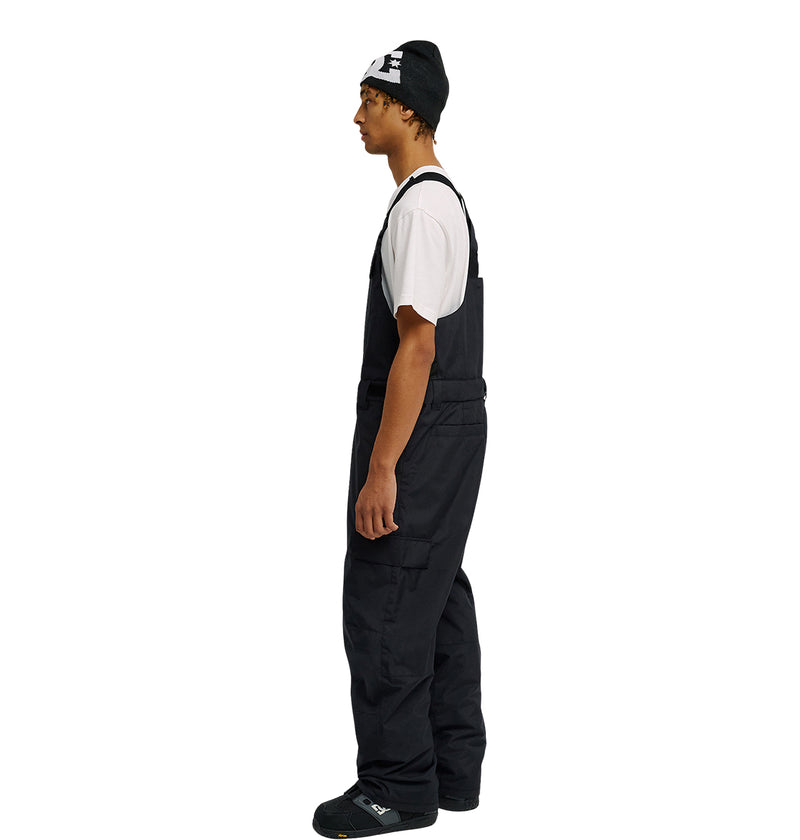 A person stands in profile against a plain white background, wearing a black beanie, white t-shirt, black shoes, and the DC Shoes Docile Bib 10K black insulated bib overalls.