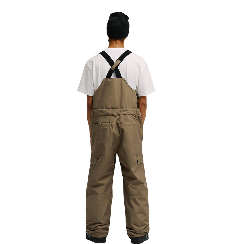 A person wearing the DC Shoes Docile Bib 10K brown overalls with breathable protection, a white t-shirt, and a black beanie stands facing away from the camera against a white background.