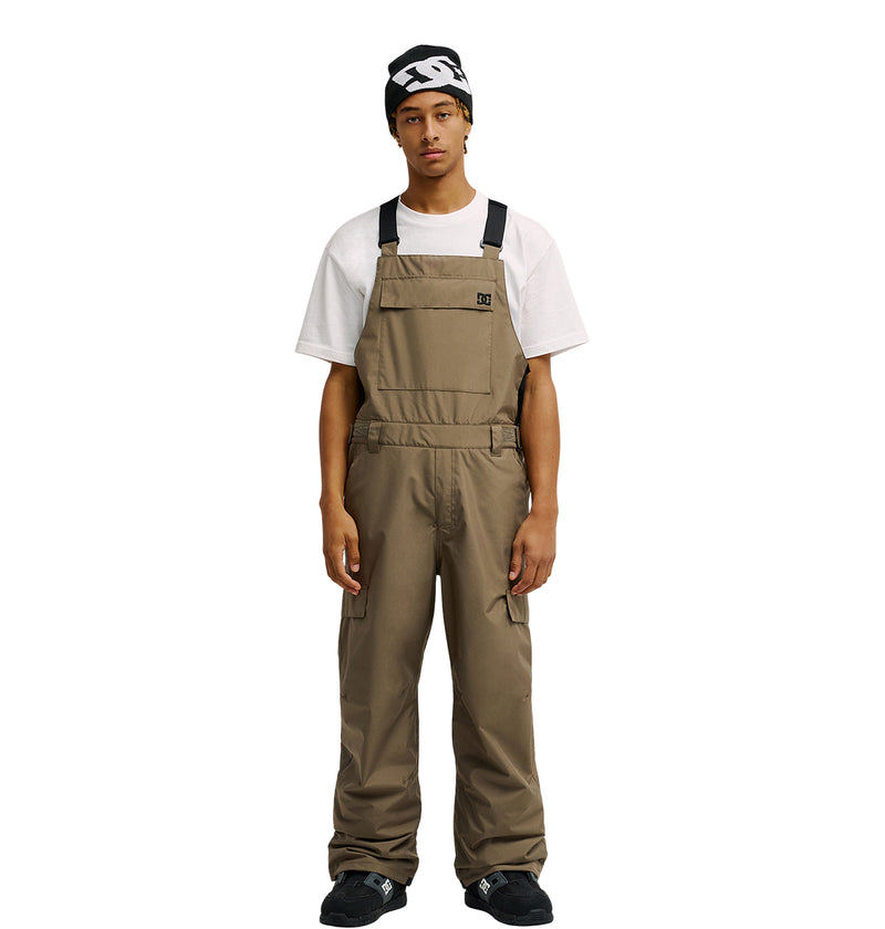 A person wearing a white T-shirt, the DC Shoes Docile Bib 10K tan overalls designed with breathable protection, a black and white beanie, and black shoes stands facing forward against a white background.