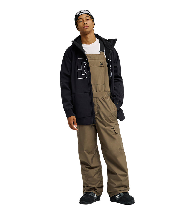 A person stands wearing a black jacket with a large 05 on the front, paired with DC Shoes’ Docile Bib 10K brown snow bib overalls for breathable protection, a white shirt, black shoes, and a black-and-white beanie.