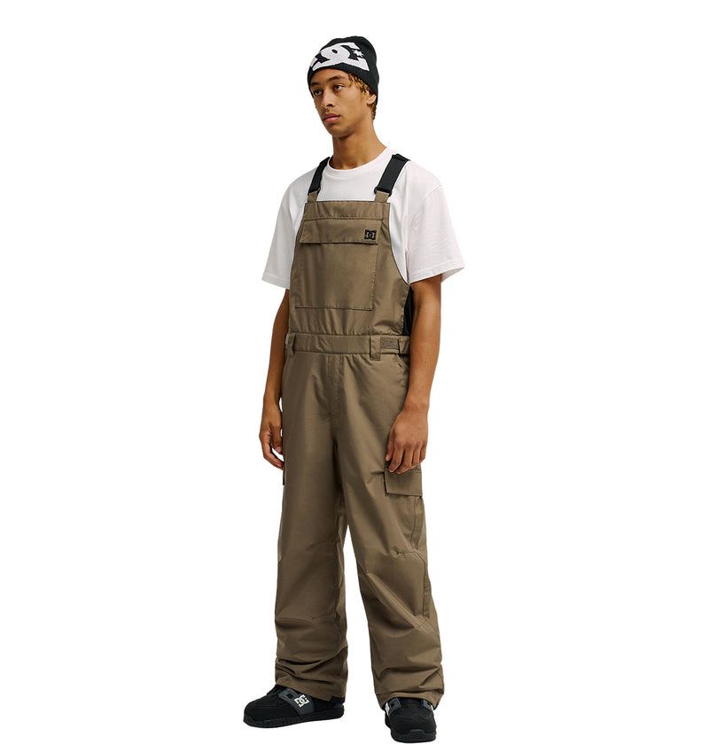 A young man wearing a black beanie, white T-shirt, and the DC Shoes Docile Bib 10K loose-fitting brown overalls with black straps, which feature breathable protection, stands against a plain white background.
