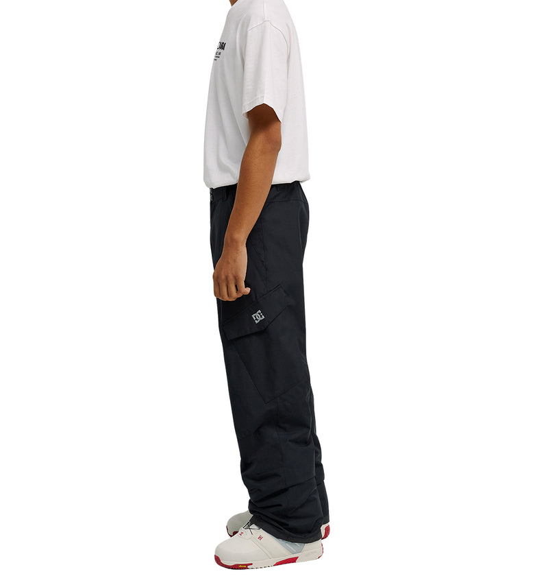 A person stands in profile against a plain white background, wearing a white t-shirt, the DC Shoes Banshee Pants—loose black waterproof cargo pants—and white sneakers with red accents.