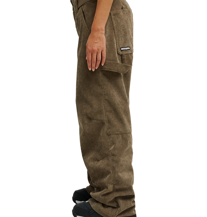 A person stands in profile wearing DC Shoes’ Snow Carpenter Pant 10K in brown, featuring a patch on the pocket. They pair the pants with black shoes, their left hand resting by their side. The plain white background emphasizes the advanced weather protection built into the design.