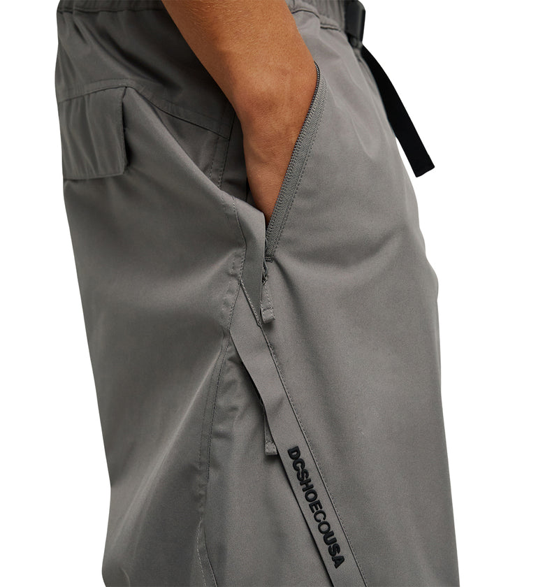 A person wearing DC Shoes Smoke Pants 10K in gray, featuring a breathable design, a black side zipper pocket, and “DSHO DECOUSA” text detailing, rests their hand in the pocket.
