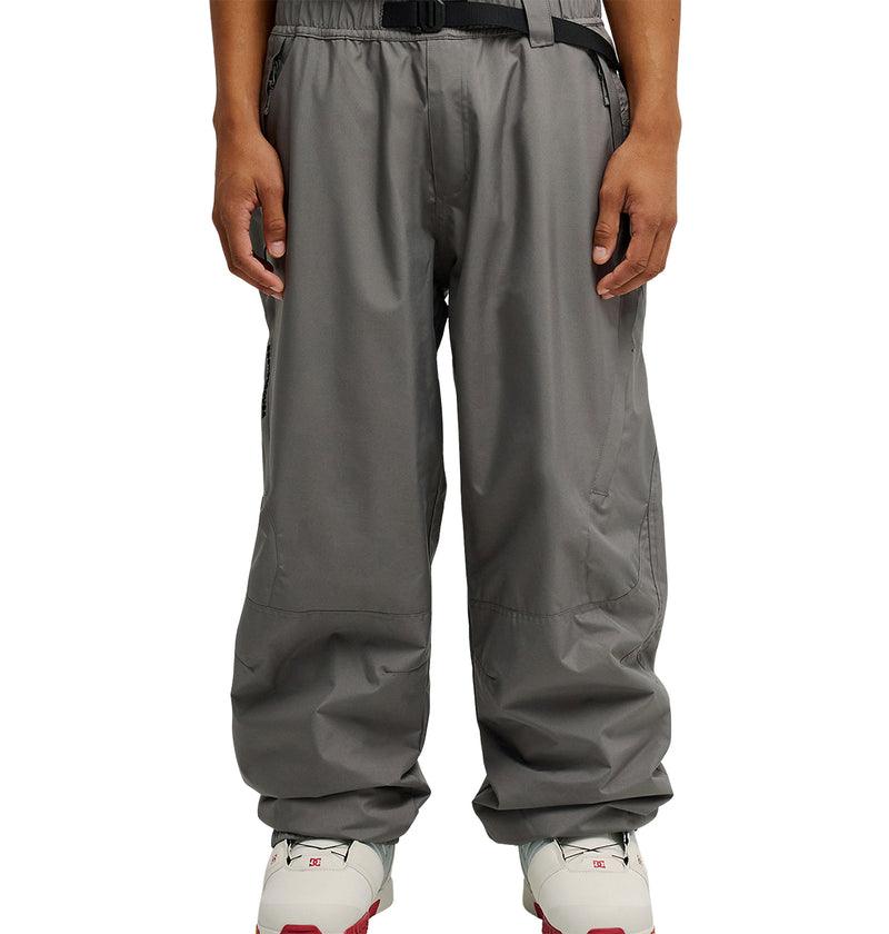A person is wearing DC Shoes’ Smoke Pants 10K, loose-fitting gray pants designed for breathable protection, paired with white sneakers featuring red accents, standing with arms relaxed at their sides.