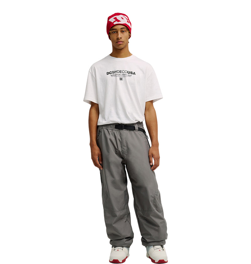 A young person wearing a red beanie, white graphic T-shirt, DC Shoes Smoke Pants 10K in gray with a black belt, and white sneakers stands facing the camera against a plain white background.