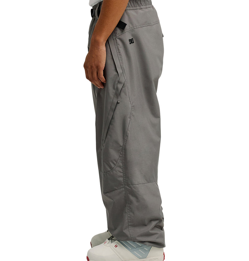A person is standing sideways, showcasing the lower half of their body dressed in DC Shoes Smoke Pants 10K—loose-fitting gray pants designed for breathable protection and equipped with practical pockets and a small logo near the pocket. They are also wearing white sneakers.
