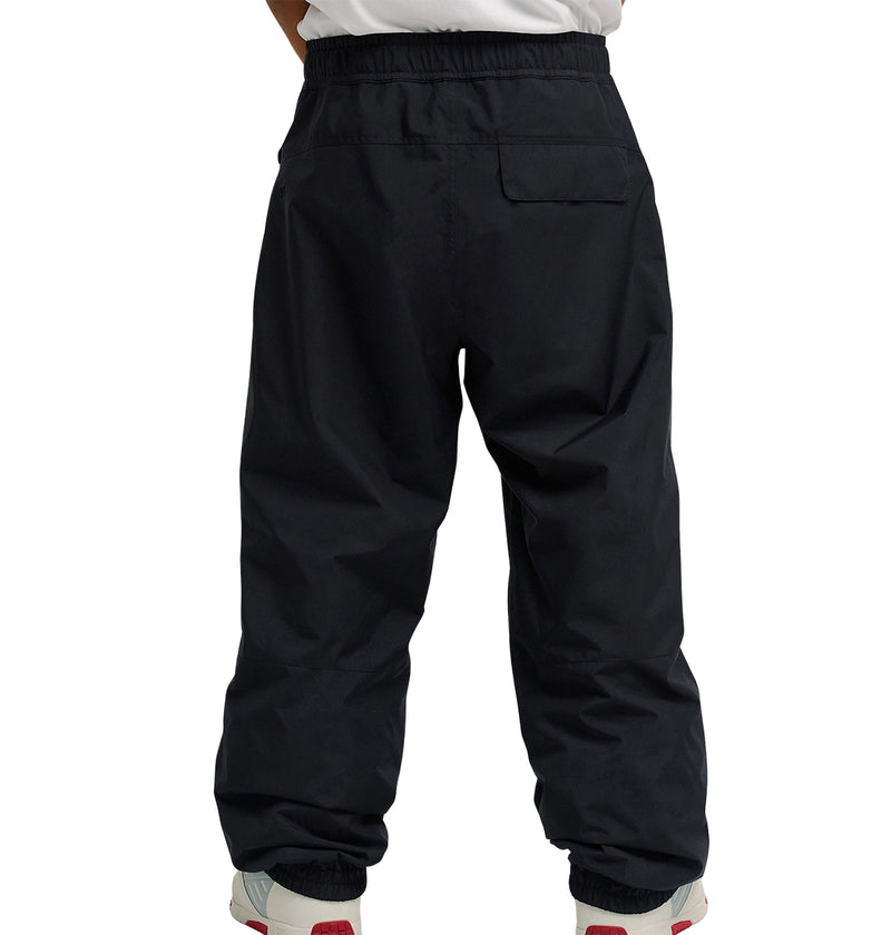 A person wearing DC Shoes Smoke Pants 10K—loose-fitting black pants designed for breathable protection—with white sneakers featuring red accents, shown from the back against a plain white background.