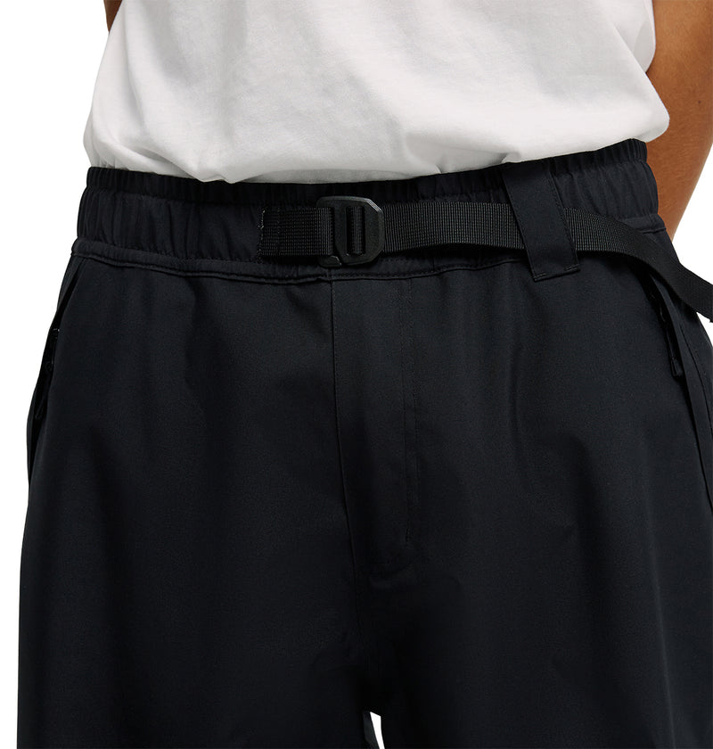 A person wearing DC Shoes Smoke Pants 10K—black pants with a built-in adjustable belt—and a white shirt, shown from the waist down. The pants provide breathable protection for added comfort during any activity.