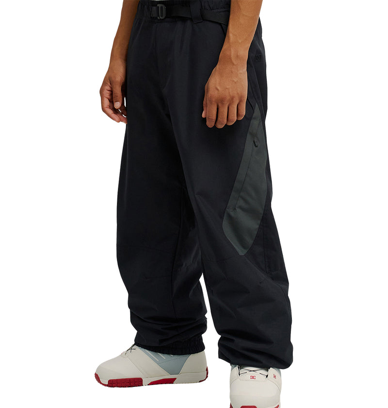 A person is wearing the DC Shoes Smoke Pants 10K in black and dark gray, featuring a side pocket and a belted waist, paired with white sneakers with red soles, while standing against a plain white background. The Smoke Pants 10K offer breathable protection and are engineered by DC Shoes for excellent snow performance, combining style with functionality.