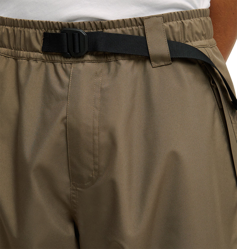 Close-up of a person wearing DC Shoes Smoke Pants 10K—khaki, breathable pants featuring an integrated black belt, elastic waistband, and belt loops, paired with a white shirt tucked in.