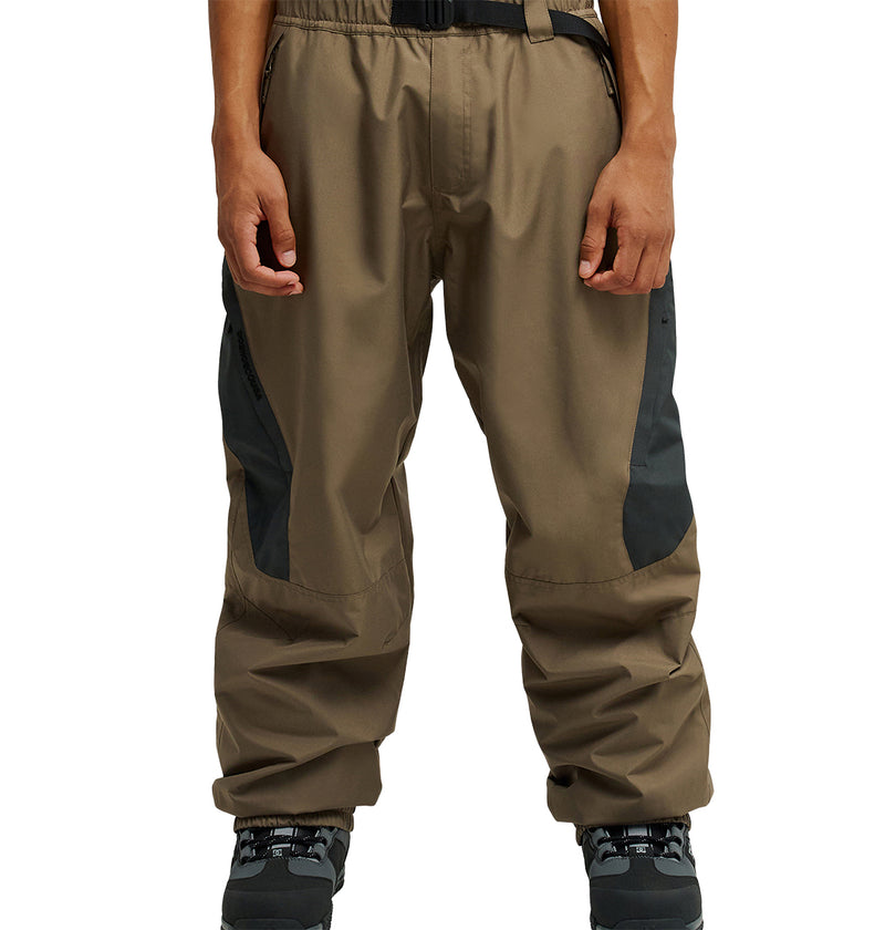A person is shown from the waist down wearing DC Shoes Smoke Pants 10K—loose-fitting brown snow pants featuring black knee patches, a black belt, and black shoes; the image highlights the breathable design.