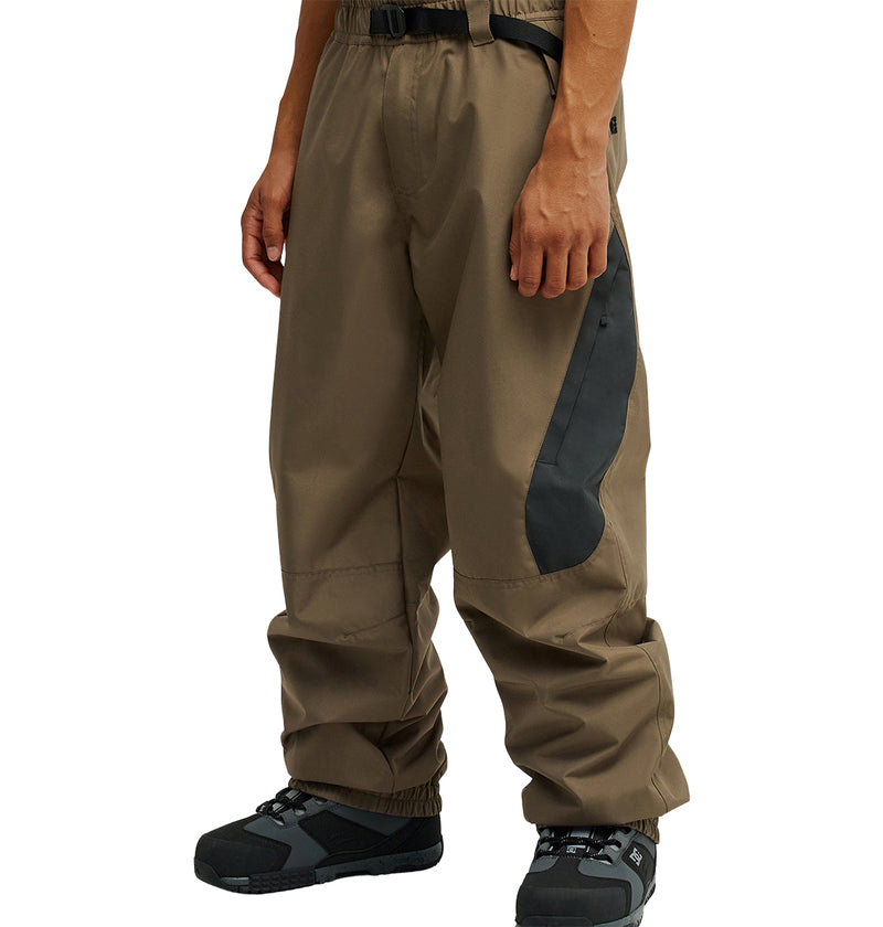 A person shown from the waist down wearing DC Shoes’ Smoke Pants 10K—loose brown waterproof outdoor pants featuring black side patches—paired with black hiking shoes.