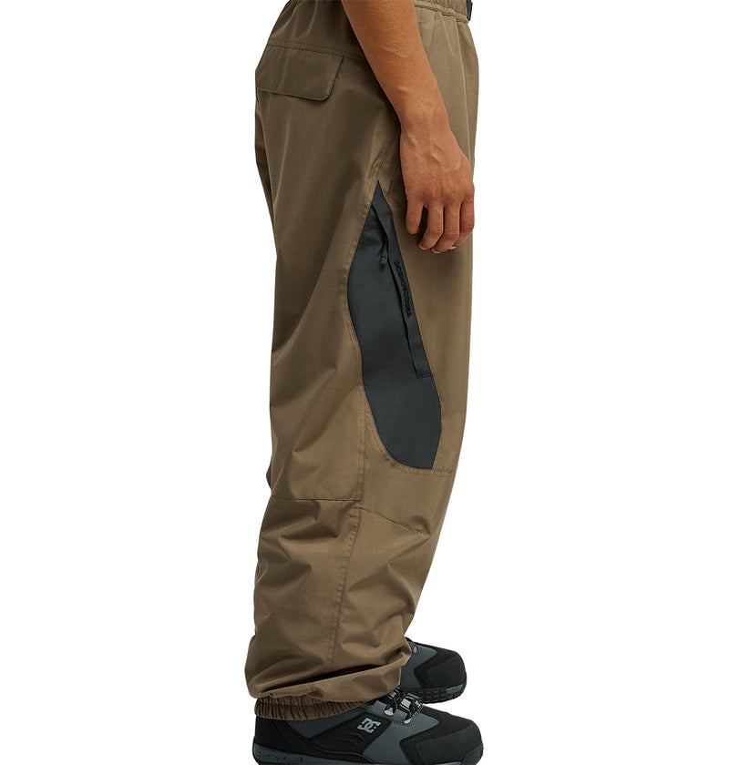 A person wearing DC Shoes Smoke Pants 10K in brown, featuring a breathable design and a black side zipper pocket, paired with black sneakers and shown in a side profile view.