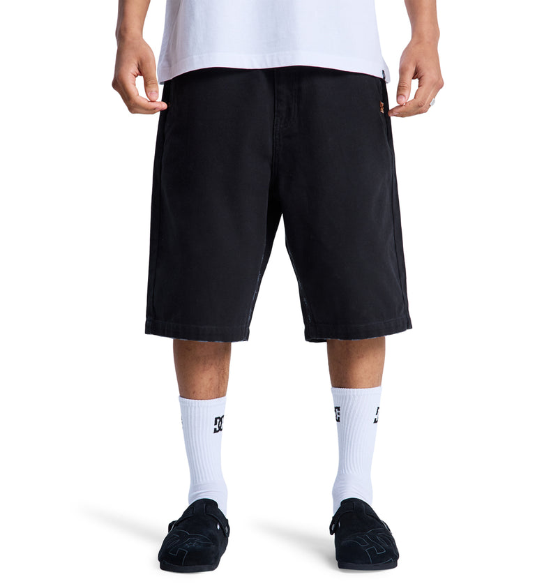 A man is seen from the waist down wearing Reebok Stressed 5 Pocket black knee-length shorts, a white t-shirt, white DC socks, and black slip-on shoes against a white background.