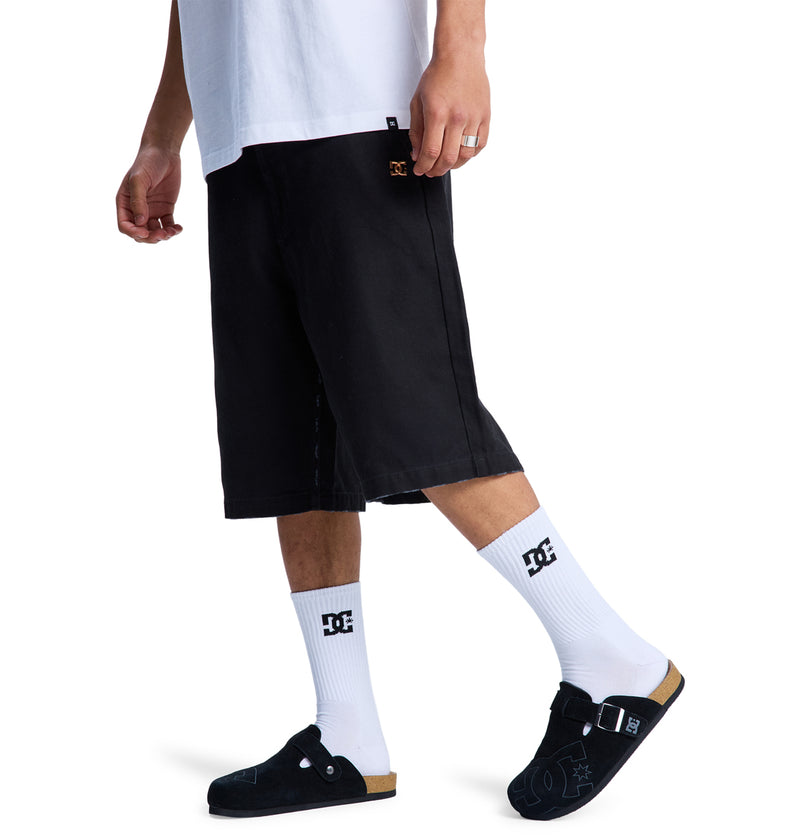 A person is seen from the waist down walking against a white background, wearing Reebok Stressed 5 Pocket black knee-length shorts, a white t-shirt, white DC logo socks, and black slip-on sandals.