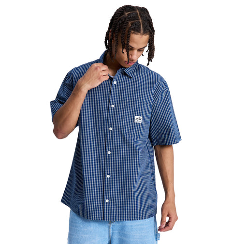 A person wears the Reebok Game On Ss, a blue and white checkered short-sleeve button-up shirt with a chest patch, paired with light blue shorts, looking down while touching their collar.