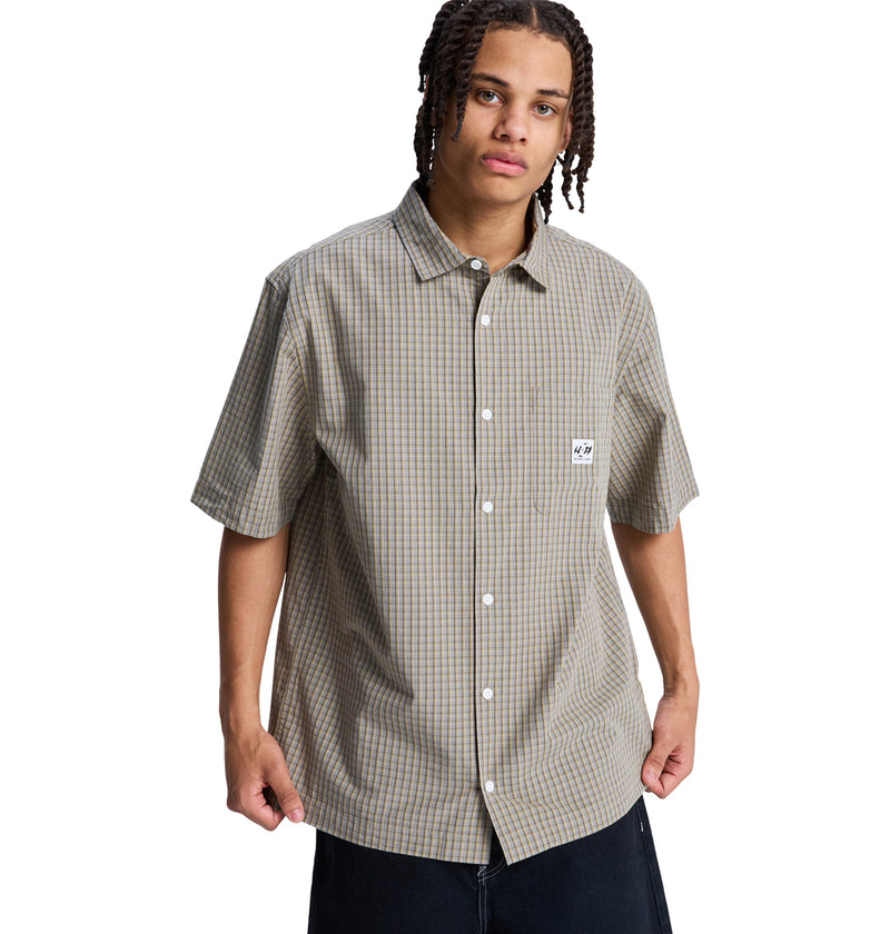 A person with medium-length braided hair wears the Reebok Game On Ss short-sleeved plaid button-up shirt, featuring a small white chest patch and dark pants, standing against a plain white background.