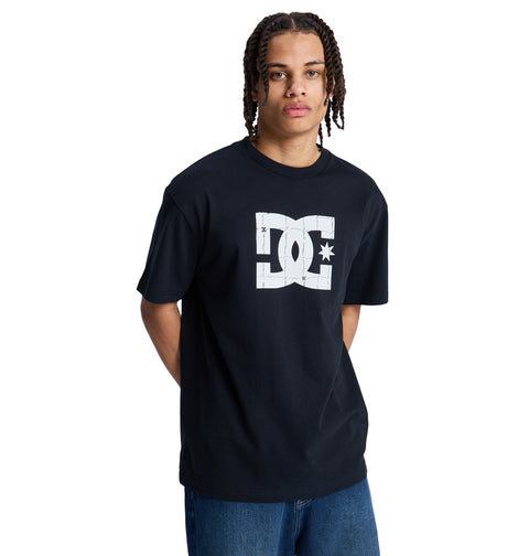 A person with medium-length braids wears a black Reebok DC Star Filled Hss t-shirt with a large white logo on the front and blue jeans, standing against a plain white background.