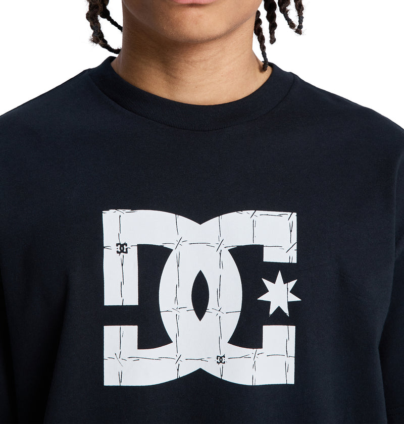 A person wears a black Reebok DC Star Filled Hss shirt with a large white logo on the front featuring a barbed wire design. Only the lower half of their face is visible.