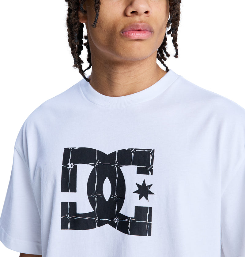 A person with braided hair wears a white Reebok DC Star Filled Hss T-shirt, featuring a large black barbed wire logo across the chest. The image is cropped to show only their upper torso and lower face.