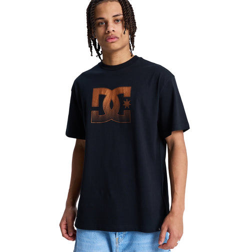 A person with braided hair wears a black Reebok DC Star Oxidized Hss t-shirt with a large brown logo on the front and light blue jeans, standing against a white background.