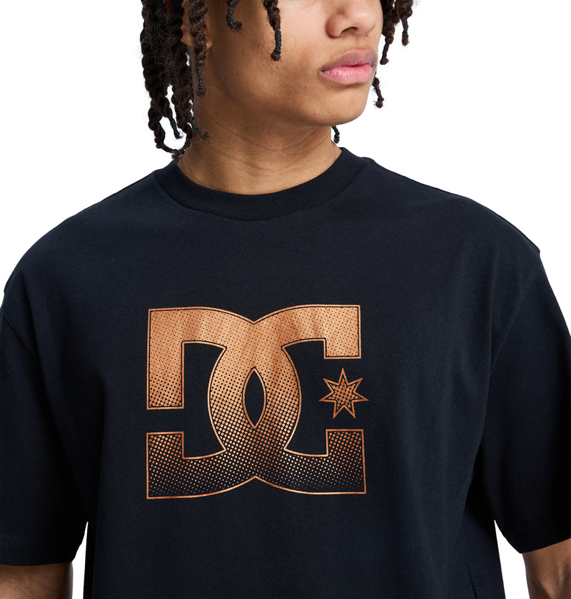 A person with curly hair wears the Reebok DC Star Oxidized Hss black t-shirt, featuring a large orange stylized DC logo with a star on the front. Only their upper body is visible as they look to the side.